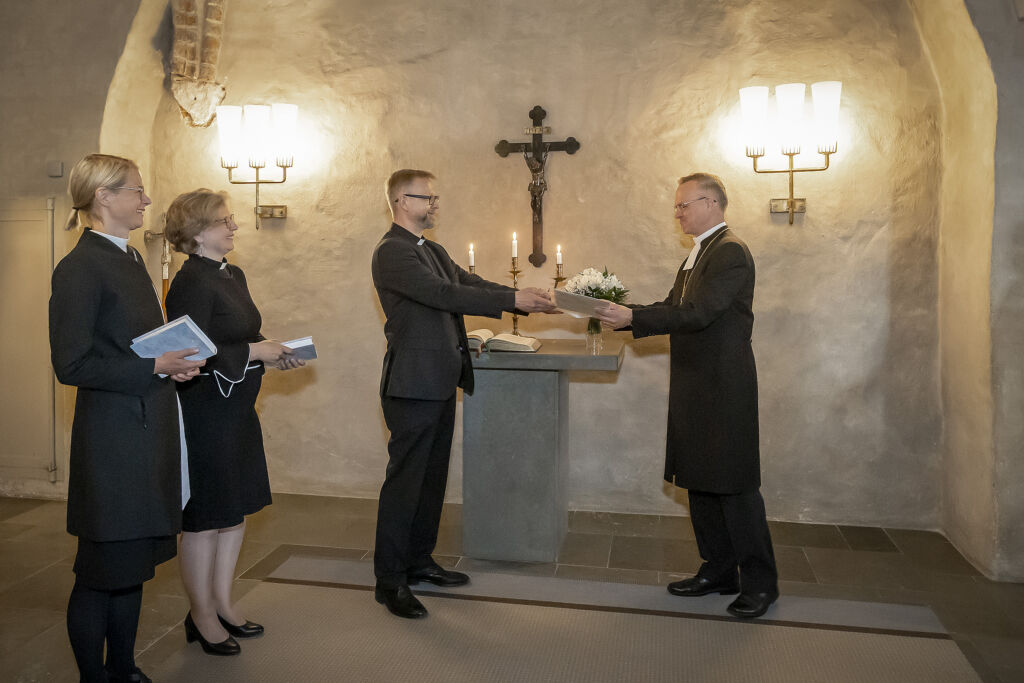 Neljä ihmistä seisoo alttarin edessä. Yksi ihmisistä ojentaa toiselle kirjan.