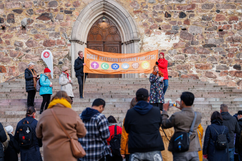 Mari Leppänen puhuu kirkon portailla Uskot yhteiskuntarauhan puolesta -bannerin edessä.